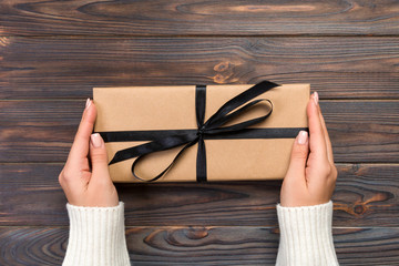 Woman hands give wrapped valentine or other holiday handmade present in paper with black ribbon. Present box, decoration of gift on dark wooden table, top view with copy space