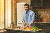 Fototapeta  - It's so delicious! Casual happy young man preparing salad at home in loft kitchen and smiling, using laptop.