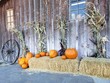 An autumn decorative harvest display of pumpkins, hay bails, corn stalks, and a wagon wheel against a wooden barn. 