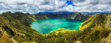 Fototapeta  - A bird's eye panoramic view of the bright green volcanic Quilotoa Lake in Ecuador with lots of white and grey clouds in a blue sky and green brush on the side of the caldera