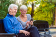 © lordn - Smiling senior active couple sitting on the bench looking at smartphone