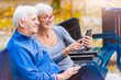 © lordn - Smiling senior active couple sitting on the bench looking at smartphone