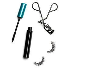 An open bottle of mascara with a black body and blue-green cap, black steel eyelash curler, and 2 long dramatic false eyelashes arranged on the left side on white background shot with studio light