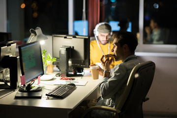 Group of people working together at night, focus on man eating snacks at workplace