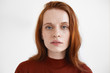 Gorgeous blue eyed red haired young European lady in maroon turtleneck looking at camera with calm facial expression, posing isolated against blank studio wall background with copyspace for your text
