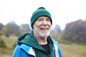 Portrait of senior countyman having walk in rural area. Cheerful elderly farmer spending time outoors. Mature retired male with beard walking in wild nature, looking at camera with happy smile