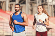 © proimagecontent - Sportish pretty blonde long haired woman in white t-shirt and multicultural handsome bearded man in blue shirt jogging at the football stadium outdoor, healthy lifestyle and people concept