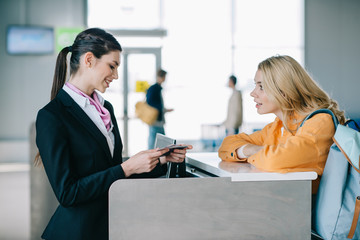 side view of smiling airport worker checking documents of young female traveler at check-in desk