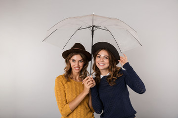 Wall Mural - Image of two joyful women wearing hats standing under umbrella, isolated over gray background