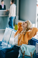 Wall Mural - attractive happy girl with hands behind head sitting and looking up in airport terminal