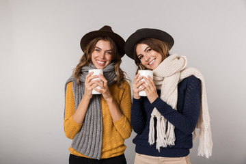 Sticker - Image of two gorgeous women wearing hats and scarfs drinking hot tea from cups, isolated over gray background