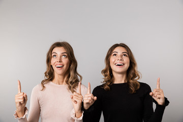 Wall Mural - Image of two lovely girls smiling and pointing fingers upward together, isolated over gray background