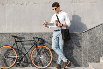 Wall Mural - Photo of young man 30s wearing sunglasses, drinking takeaway coffee and reading newspaper, while standing with bicycle along wall outdoor