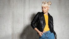 Stylish Woman Wearing Leather Jacket. Young Blond-haired Female Posing In Studio On Cement Background. Modern Fashion Concept.