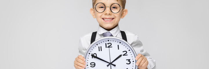 A charming boy in a white shirt, suspenders, a tie and light jeans stands on a gray background. The boy is holding a watch