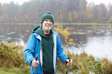 Outdoor Shot Of Happy Senior Man In Warm Clothes Standing With Nordic Walking Poles In Colorful Autumn Park With Lake In Background. Healthy Lifestyle, Activity, Wellness And Fitness Concept