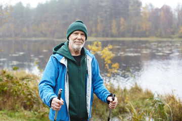 Outdoor shot of happy senior man in warm clothes standing with nordic walking poles in colorful autumn park with lake in background. Healthy lifestyle, activity, wellness and fitness concept