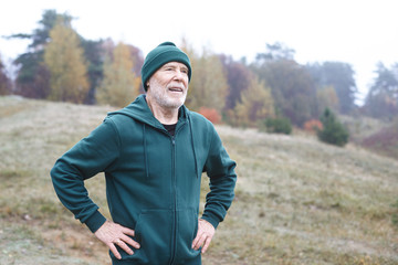 Outdoor shot of elderly athletic unshaven man in green hat and hoodie holding hands on waist, catching breath after cardio running workout, having tired look. Healthy and active lifestyle at any age