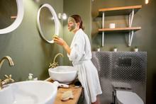 Young Woman In Bathrobe Applying Green Mask On Her Face Standing Near The Mirror In The Modern Green Bathroom