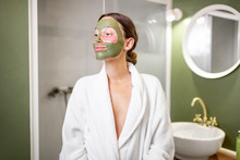 Portrait Of A Young Woman In Bathrobe With Facial Green Mask And Patches Under Her Eyes In The Bathroom