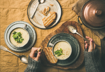 Wall Mural - Autumn or Winter home dinner. Flat-lay of Fall warming celery cream soup in cups and female hands in sweater over linen tablecloth, top view. Comfort food, vegetarian, healthy and slow food concept