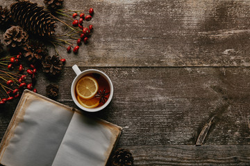 Wall Mural - flat lay with red holly berries, blank notebook, cup of tea and pine cones on wooden tabletop