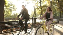 Close Up Of Caucasian Young Couple Or Friends Riding Their Bikes In The Empty City Park Or Boulevard In Summertime. People, Leisure And Lifestyle Concept. Green Trees Around, Sun Shines On The