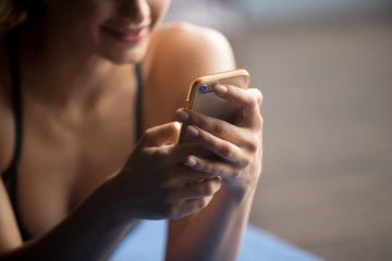 Young attractive woman after practicing yoga, relaxing on yoga mat, using app, making a call, texting, smiling and enjoying a rest, looking at smartphone, messaging, indoor close up of hands and phone