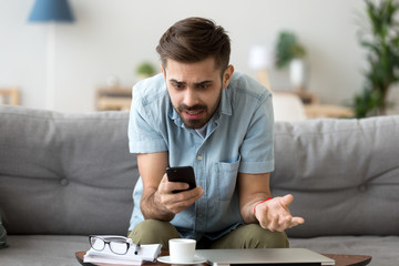 Confused millennial man sitting alone on couch in living room at home looking at smartphone screen with irritation and indignance. Annoyed male received bad news, device gadget broken or dead concept