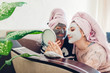 © maryviolet - Mother and her adult daughter looking at mirror with facial masks applied. Women chilling and having fun