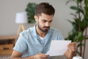 Frustrated millennial man sitting at the desk in office holding reading notification about debt or not recruited. Stressed student holding letter about scholarship refusal expulsion from high school