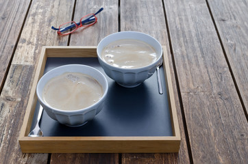 Two cups of milk and coffee in a tray on a wooden table. In the background red/blue glasses.