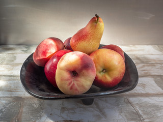 Ripe nectarines, pears, apples in a black dish are on a light wooden table.