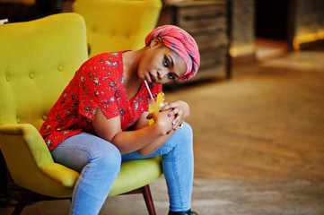 Wall Mural - Stylish african woman in red shirt and hat posed indoor cafe and drinking pineapple lemonade.