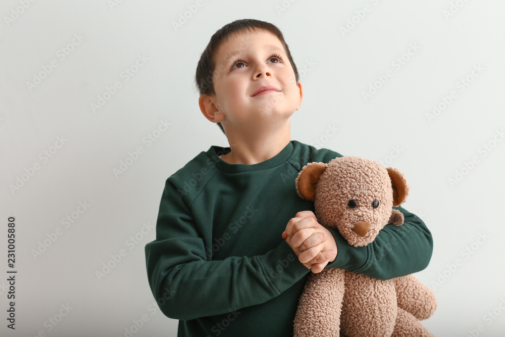 Little boy with teddy bear praying on light background
