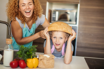 Beautiful mother and cute little daughter having fun in the kitchen while preparing to cook muffins in the bowl. Happy girl wears the bowl on head and smiling.