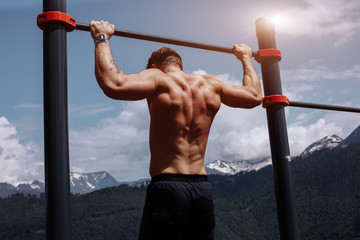 Naked torso athlete man training pull ups in amazing nature mountains landscape. Strength training fit male working out exercising outdoors in summer doing pull-ups and chin-ups on horizontal bar.