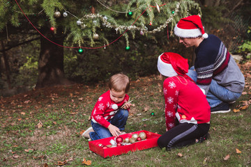 Merry Christmas and Happy Holidays. Father in red Christmas hat and two daughters in red sweaters decorating the Christmas tree outdoor in the yard of the house before holidays