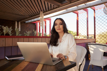 Young bisnesswoman copywriter keyboarding information  while sitting in summer terracecafeteria. Kazakh student looking at computer while writing text message to someone using wireless 4G wifi