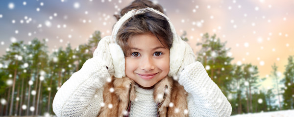 Poster - winter, people and christmas concept - happy little girl wearing earmuffs over snowy forest background