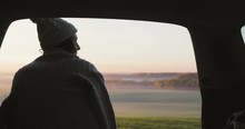 Young woman admiring the sunrise in trunk of car. Girl dressed in woolen socks drinking hot coffee against backdrop of forest panorama in fog . View from inside 4k video