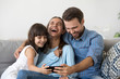 © fizkes - Smiling happy multi-ethnic family sitting on couch in living room at home. Mother father and little daughter laughing using mobile phone watching funny videos online have fun looking humorous program