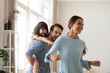 © fizkes - Young happy father laughing carrying on back piggybacking little daughter catching mum playing with diverse family having fun together. Kid enjoying active game with parents in living room at home