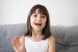 Small girl sitting on couch alone at home. Head shot portrait little adorable daughter waving hand looking at camera smile saying hello or goodbye. First acquaintance and online communication concept