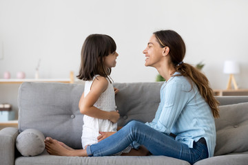 Attractive woman and little girl sitting on comfortable couch at home. Young mother talking communicates with small adorable daughter. Best friends happy motherhood weekend together with kid concept