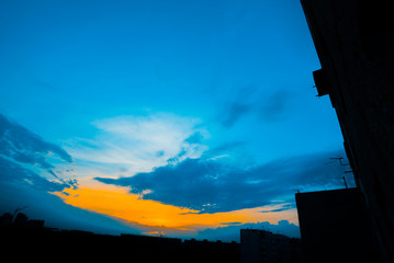 Wall Mural - Atmospheric blue cloudy sky behind silhouettes of city buildings. Cobalt and orange background of sunrise with dense clouds and bright yellow sunny light for copy space. Cyan heaven above clouds.