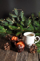 Christmas Breakfast of two cupcakes and a Cup of hot coffee on a wooden table, on the background of fir branches with lights.