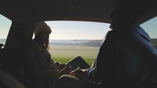 Personal perspective of couple lying in car's trunk, mountain landscape and admiring sunrise . Lovers alking and drinking hot coffee against backdrop of forest panorama in fog . View from inside