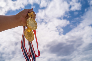 Winner hand raised holding two gold medals with Thai ribbon against blue sky. Golden medals is award for highest achievement for sport or business. Success Awards concept