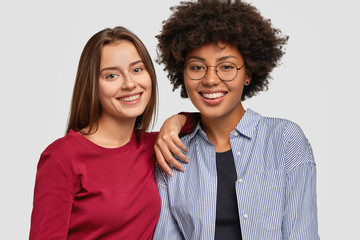 Multiethnic women with cheerful expressions, have positive smiles, pose together, stand against white background. Diverse multicultural females have friendly relationships. People and joy concept